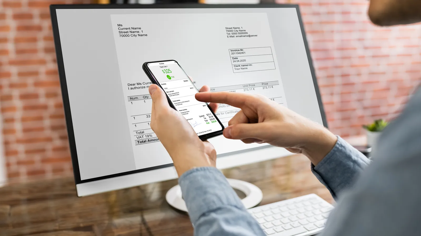 A hand paying an invoice on a phone in front of a computer screen