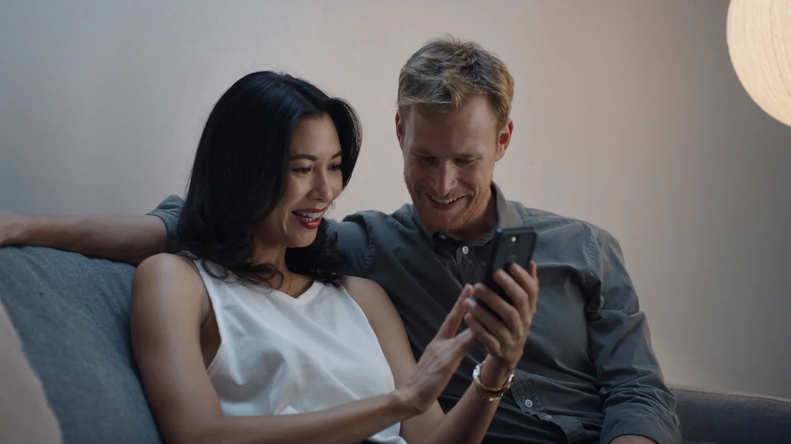 An engaged couple on a sofa in the evening, smiling at a phone together