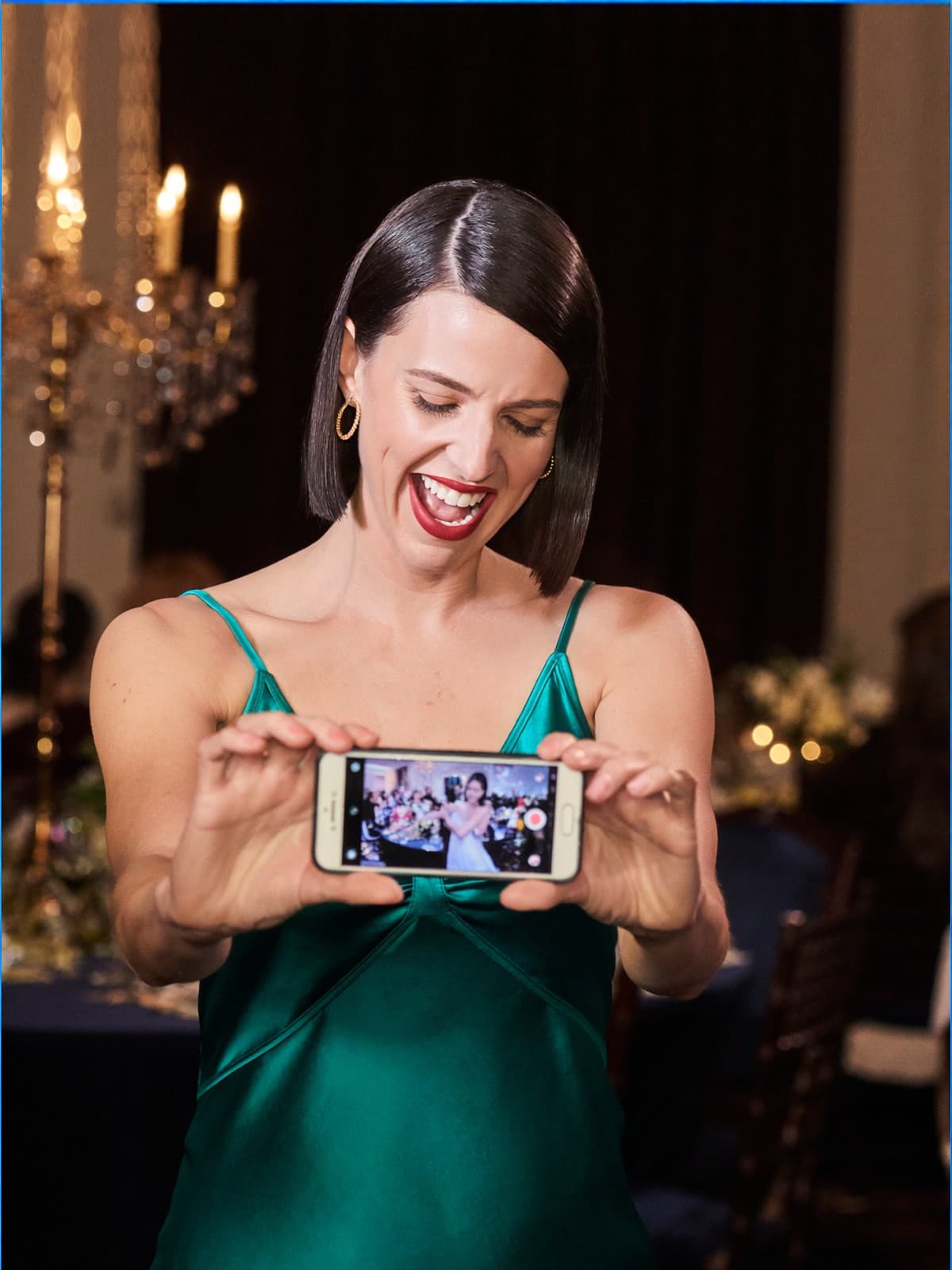 A wedding guest in emerald silk laughing as she films the first dance on her phone
